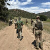 A group of individuals in tactical gear walk along a dirt road through a mountainous area under a sunny sky with scattered clouds, likely during a field training or tracking exercise.