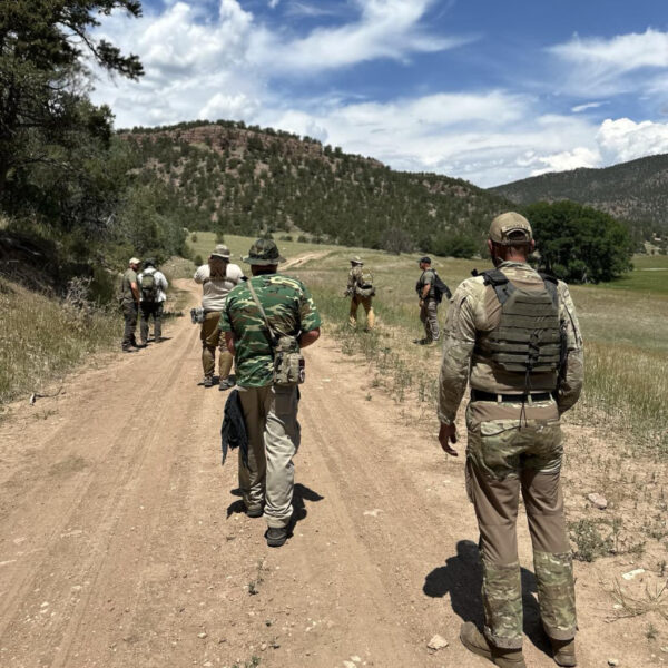 A group of individuals in tactical gear walk along a dirt road through a mountainous area under a sunny sky with scattered clouds, likely during a field training or tracking exercise.