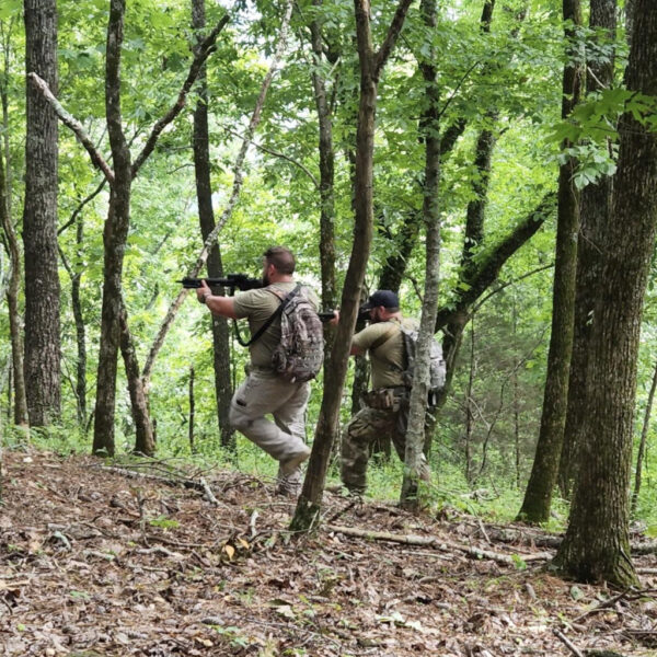 Two armed individuals moving tactically through a forest, one aiming down sights while the other follows closely behind.