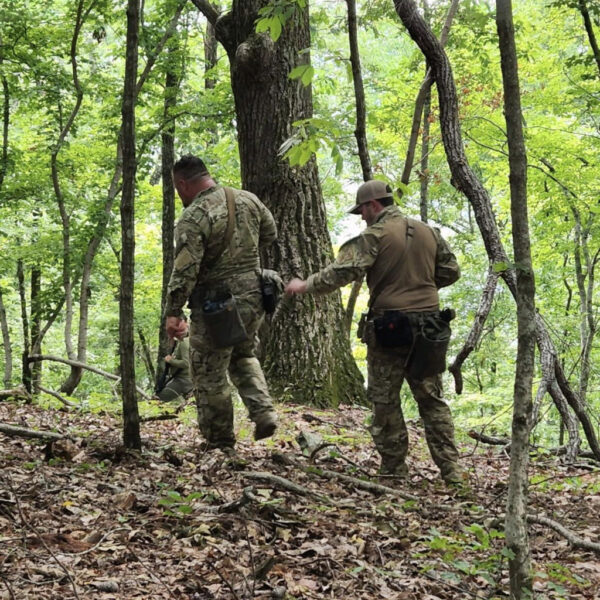Two individuals in camouflage gear tracking through a dense green forest, one pointing ahead while the other observes the ground.