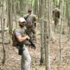 Three armed men in tactical gear move through a dense forest. The man in the foreground looks back while holding a rifle, with two others walking ahead.