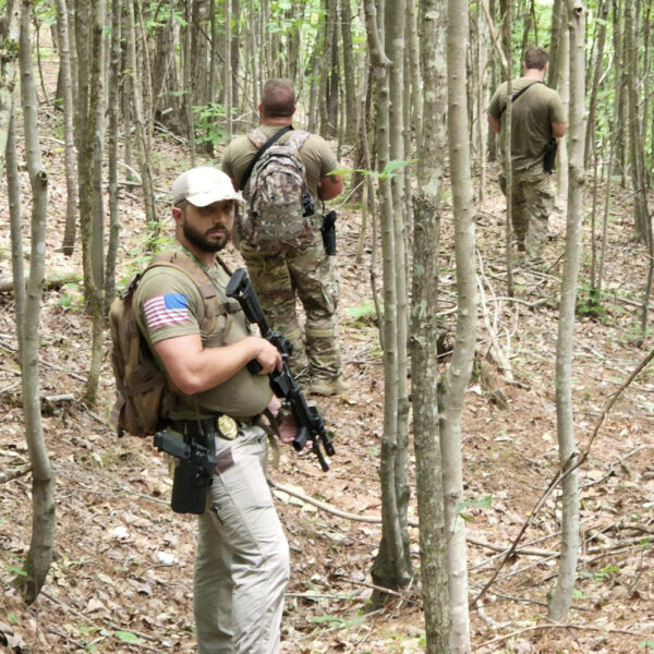 Three armed men in tactical gear move through a dense forest. The man in the foreground looks back while holding a rifle, with two others walking ahead.