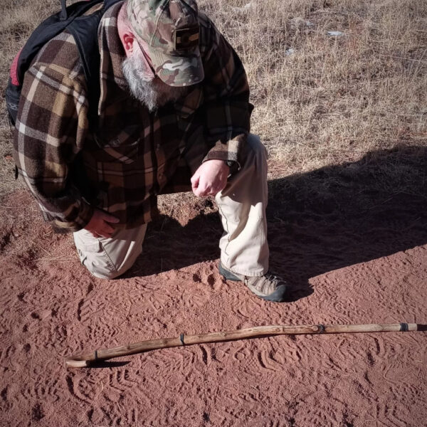 A man kneels in a field of red dirt and dry grass while studying animal tracks. He wears a camouflage hat, plaid jacket, tan pants, and hiking boots. A long wooden tracking stick is laid across the ground in front of him, aligned with the tracks.