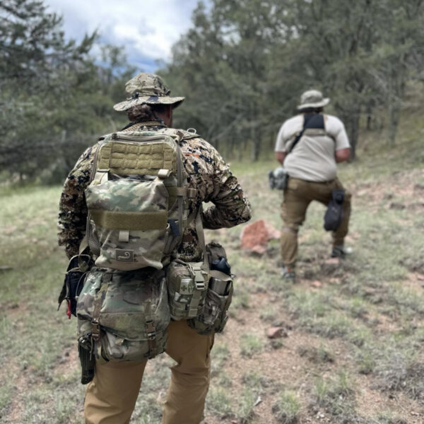 Two individuals wearing tactical backpacks and outdoor gear walk through a forested area under a cloudy sky during a field tracking or wilderness movement exercise.