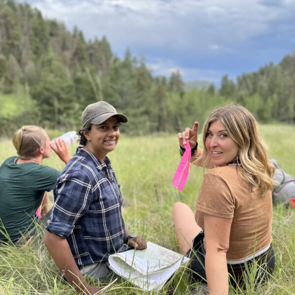 Three hikers sit in a grassy mountain clearing, one smiling with a map on their lap, another holding up a pink trail marker ribbon and flashing a peace sign, while the third drinks from a water bottle. Pine-covered hills and a moody sky stretch out behind them.