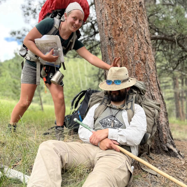 A smiling woman playfully gives bunny ears to a man resting against a tree with his hat down and hiking stick in hand. Both are in full backpacking gear in a pine forest.