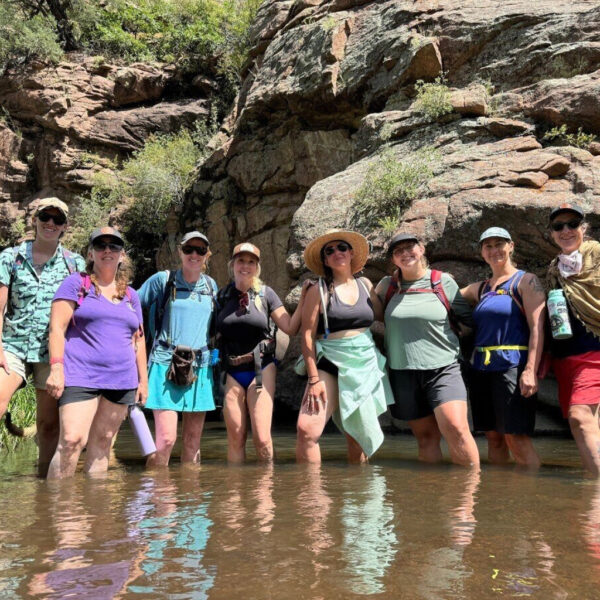 A group of women stands smiling in ankle-deep river water during the Wild Women River Retreat, surrounded by towering red canyon walls and sunlit greenery.
