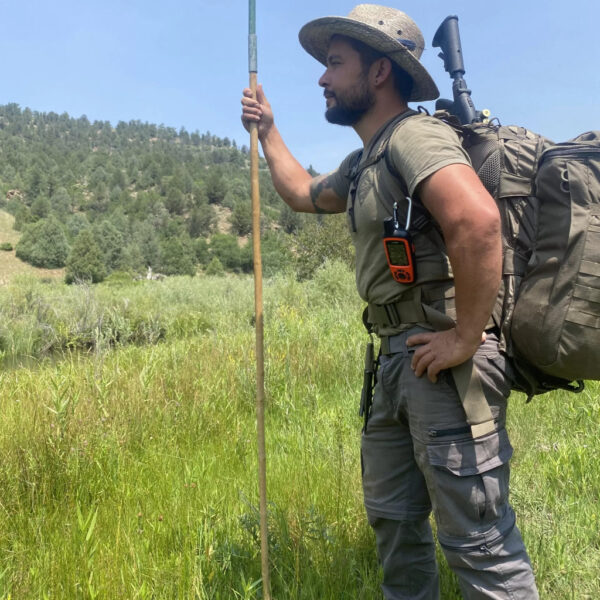 A man in tactical hiking gear stands confidently in a grassy field, holding a long wooden staff. He wears a wide-brimmed hat and carries a large backpack with outdoor tools, including a GPS device and rifle stock.