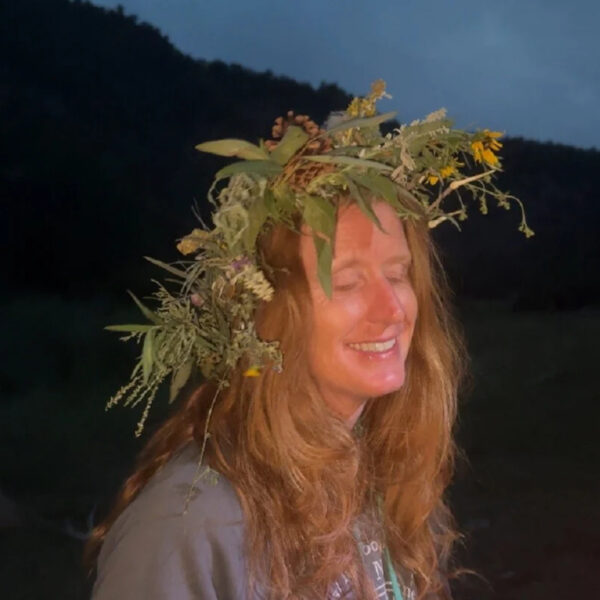 A woman smiles with her eyes closed at dusk, wearing a handmade crown of wildflowers, herbs, and forest plants during the Wild Women River Retreat.