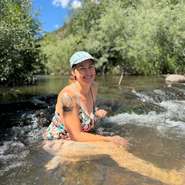A woman relaxes in a clear mountain stream, smiling under the bright sun. She wears a floral swimsuit and a patterned cap, surrounded by lush green foliage and rocky terrain in the background.