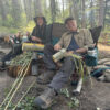 Two people sitting in camp chairs smiling and working on bark stripping projects in a wooded outdoor area. A dog is lying on the ground beside them, relaxing in the shade.