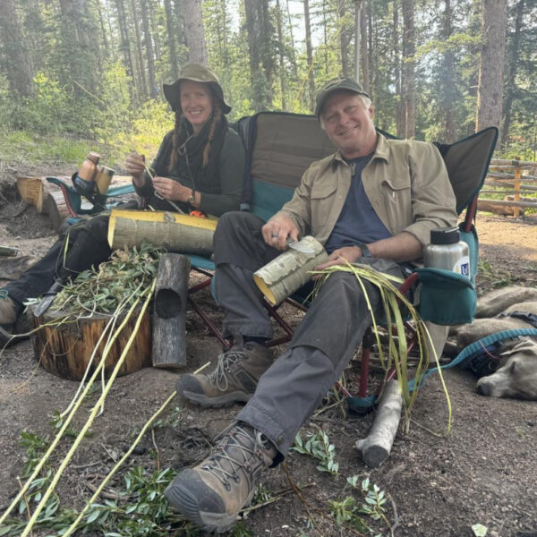 Two people sitting in camp chairs smiling and working on bark stripping projects in a wooded outdoor area. A dog is lying on the ground beside them, relaxing in the shade.