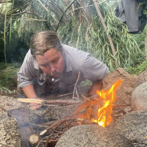 Person blowing on kindling to ignite a fire inside a natural shelter made from tree branches
