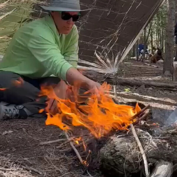 Two people building and tending a fire under a tarp shelter in a forest survival class