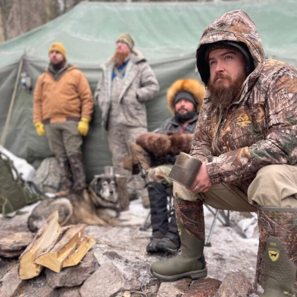 Group of men dressed in cold-weather gear gathered around a winter campsite with a green canvas tent, firewood, and a husky dog lying in the snow. One man in camouflage sits in the foreground holding a metal mug.
