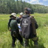 Three people standing in a sunlit mountain meadow during an ethnobotany course. One wears a “Wildcard Wilderness” hoodie with a quiver on their back. They are studying plants and taking notes, surrounded by tall grass and a forested backdrop.