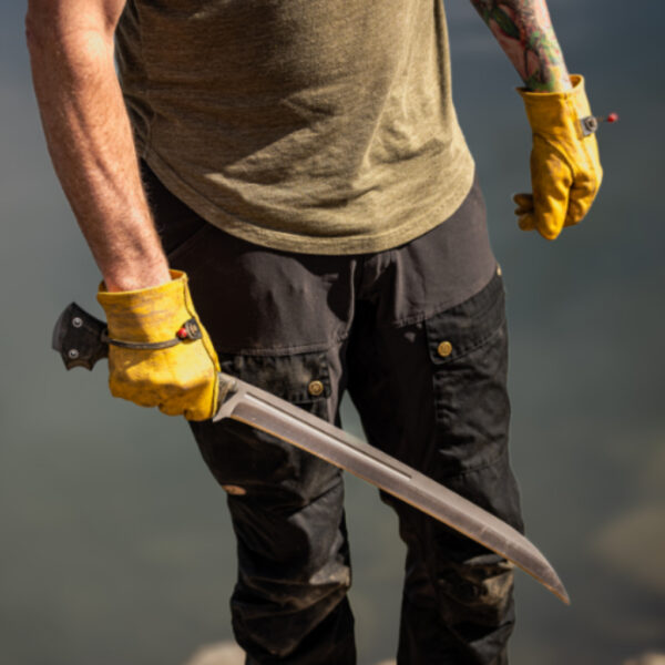 A person wearing yellow work gloves and black pants holding the Gallantry knife by their side, with the blade angled downward in front of a blurred natural background.