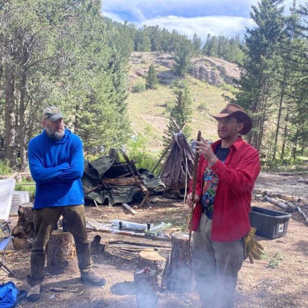 Hutch teaching about the uses of mullein near a smoky campfire in a forested mountain setting, while another participant listens attentively during a wilderness survival course at The Survival University.