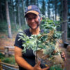 Jacks smiling with camouflage face paint holds a handmade bark basket filled with wild plants and evergreens in a forest setting. She’s wearing a dark T-shirt and a ball cap, standing near rustic fencing with tall pine trees in the background.