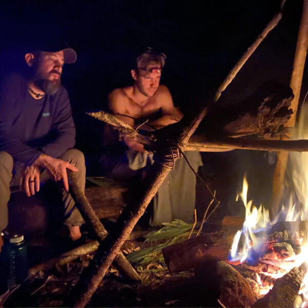 Two men sit by a glowing campfire at night in the jungle, one shirtless with a headlamp, the other in a long-sleeve shirt and cap, both focused on the flames under a primitive cooking tripod.