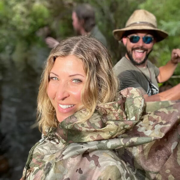 Smiling woman in camouflage rain gear takes a selfie near a stream while a man in sunglasses and a straw hat grins behind her. Another person is slightly blurred in the background, surrounded by lush greenery.