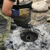 Person wearing gloves and bushcraft pants lifts a metal container from a fire pit with white ash. In the center, a tin can containing freshly rendered pine tar is visible.