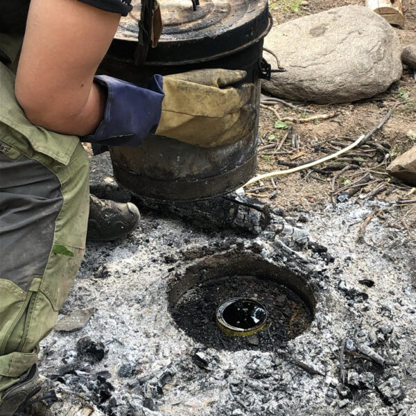 Person wearing gloves and bushcraft pants lifts a metal container from a fire pit with white ash. In the center, a tin can containing freshly rendered pine tar is visible.