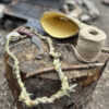 Primitive crafting tools on a tree stump: a curved knife with a wooden handle, braided plant fiber cordage, natural twine, a half gourd bowl, and a stick. A fire pit and cooking area are blurred in the background.