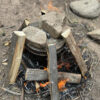 Primitive pine tar setup with a pot covered by a lid and weighted down by large rocks. The pot is surrounded by upright firewood arranged in a teepee structure, with flames and embers underneath.