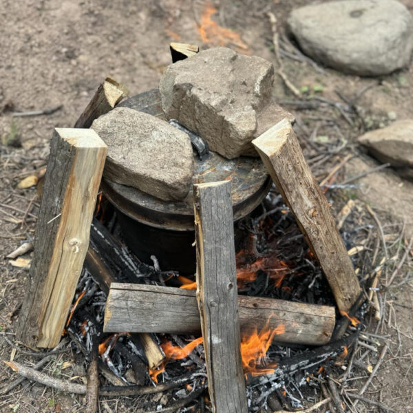 Primitive pine tar setup with a pot covered by a lid and weighted down by large rocks. The pot is surrounded by upright firewood arranged in a teepee structure, with flames and embers underneath.