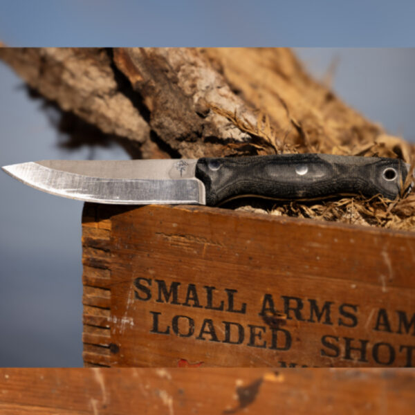 The Spirit Knife with a black micarta handle and weathered blade resting on a wooden ammo crate labeled "Small Arms Ammunition – Loaded Shot," with a rugged tree stump and blue sky in the background.