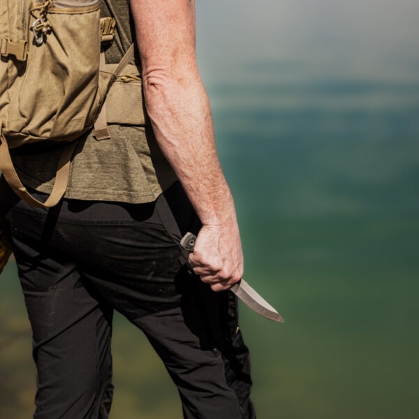 Man standing at the edge of a clear green lake, holding The Spirit Knife low in one hand. He wears black tactical pants, a green shirt, and a tan backpack with outdoor gear, facing away from the camera.