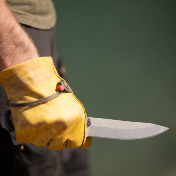 Close-up of a gloved hand holding The Spirit Knife with a clean, matte blade against a soft green outdoor background. The knife is angled forward, highlighting its edge and rugged construction.