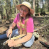 Smiling woman in a pink “Grow Gardens Not Lawns” shirt and wide-brimmed sunhat carving a stick with a knife in a forest setting during a wilderness skills workshop.