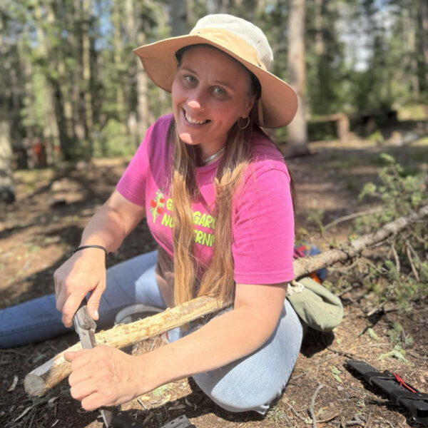 Smiling woman in a pink “Grow Gardens Not Lawns” shirt and wide-brimmed sunhat carving a stick with a knife in a forest setting during a wilderness skills workshop.