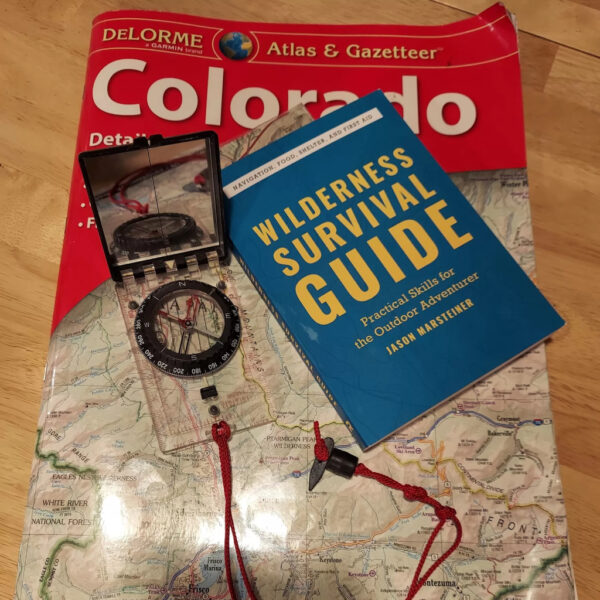 Wilderness Survival Guide book by Jason Marsteiner placed on a Colorado Atlas map alongside a baseplate compass with a red lanyard