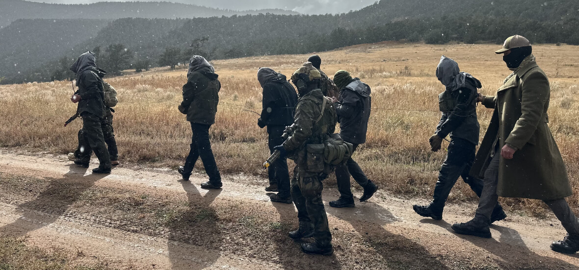SERE training participants walk in formation through a remote Colorado field with hoods over their heads, guided by an instructor during a realistic resistance exercise.