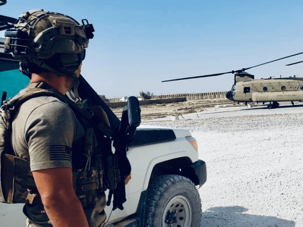 Kenny “KG” observes a military helicopter preparing for takeoff while standing beside a tactical vehicle in a desert environment.