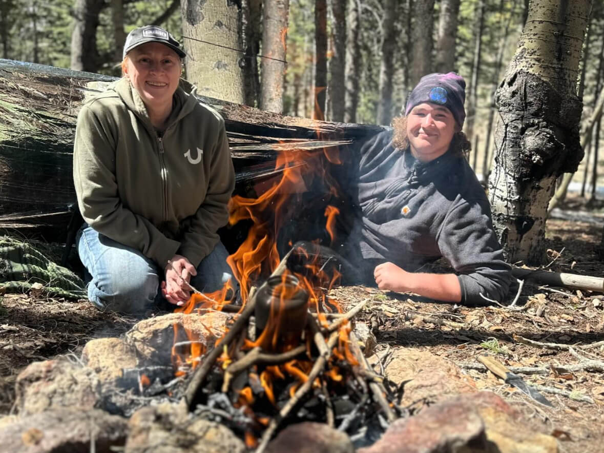 Two students sitting by a campfire beside a debris shelter during a survival training course in the Colorado mountains