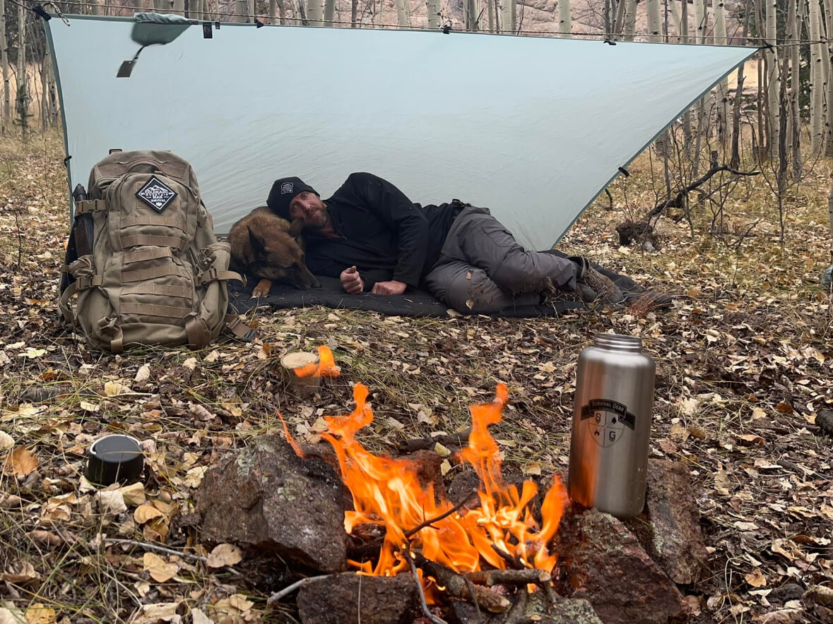 Man resting with his dog under a tarp shelter beside a campfire in the Colorado mountains during a camping course.