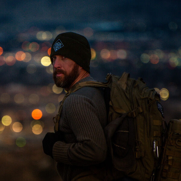 Jason Marsteiner wearing a beanie and backpack, looking over a city at night with blurred lights behind him, representing urban awareness and resilience