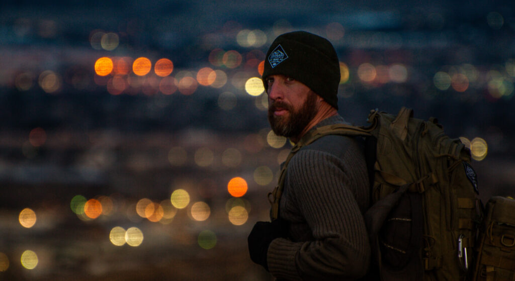Jason Marsteiner stands above a glowing city at dusk, wearing a backpack and beanie, symbolizing awareness, preparedness, and the connection between wilderness and modern survival.