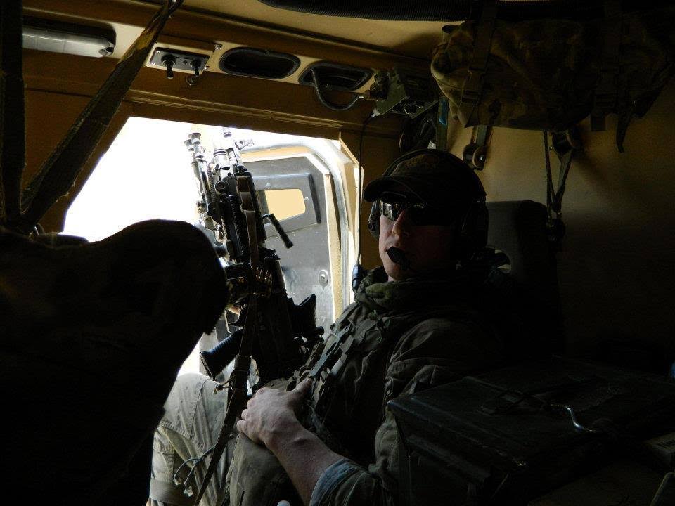 Slo seated inside a military vehicle wearing tactical gear and headset, holding a mounted weapon.