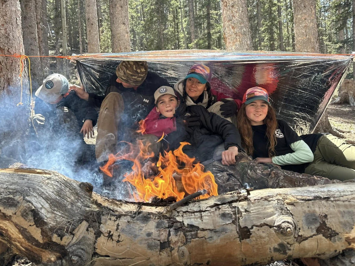 Group of students sitting under a survival shelter in the forest with a campfire burning in front of them during a wilderness training class.