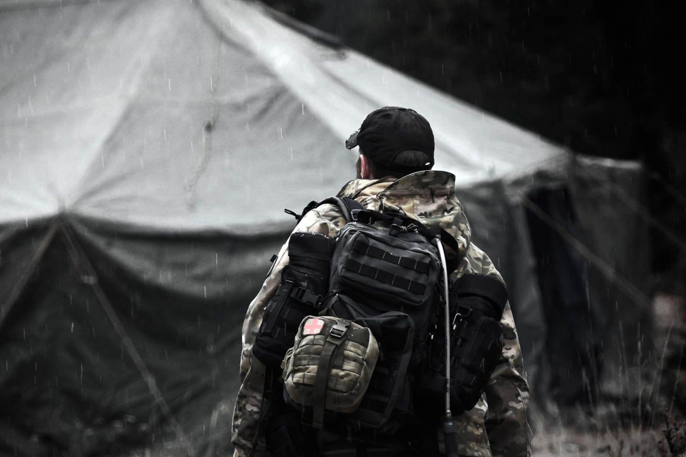 Instructor carrying a survival pack in the rain near a field tent, representing preparedness and real-world survival mindset.