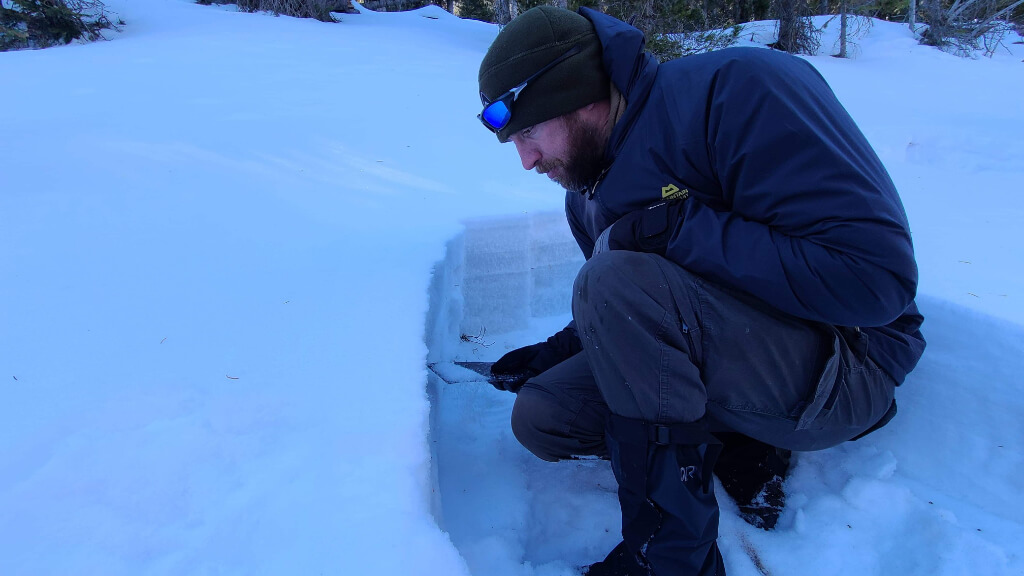 Man kneeling beside a snow pit during winter conditions, illustrating stillness and reduced movement in cold environments.