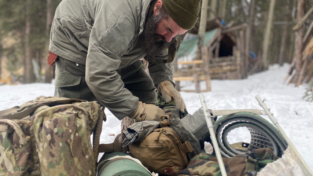 Instructor assisting a teammate with gear in snowy conditions during winter training, demonstrating hands on support and buddy care to prevent cold related injury