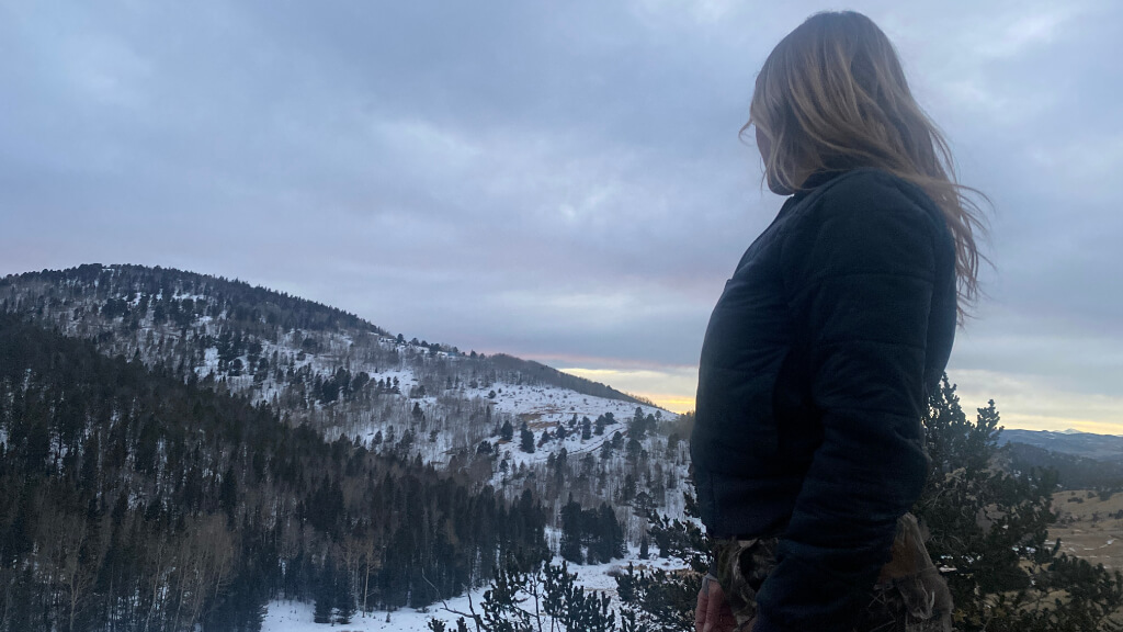 Jess Caldwell standing on a snowy hillside during winter training, looking out over mountainous terrain and forested valleys.