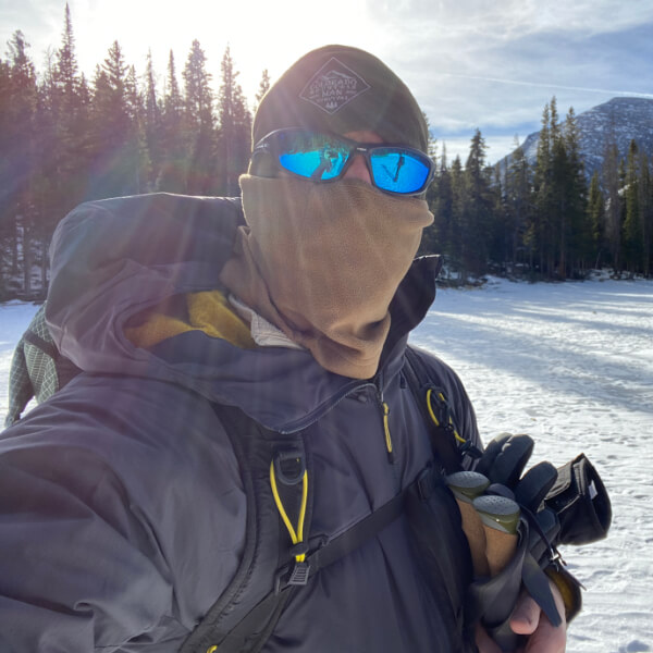 Man wearing layered winter clothing outdoors including insulated jacket, shell, gloves, face covering, and pack while standing in snowy mountain terrain