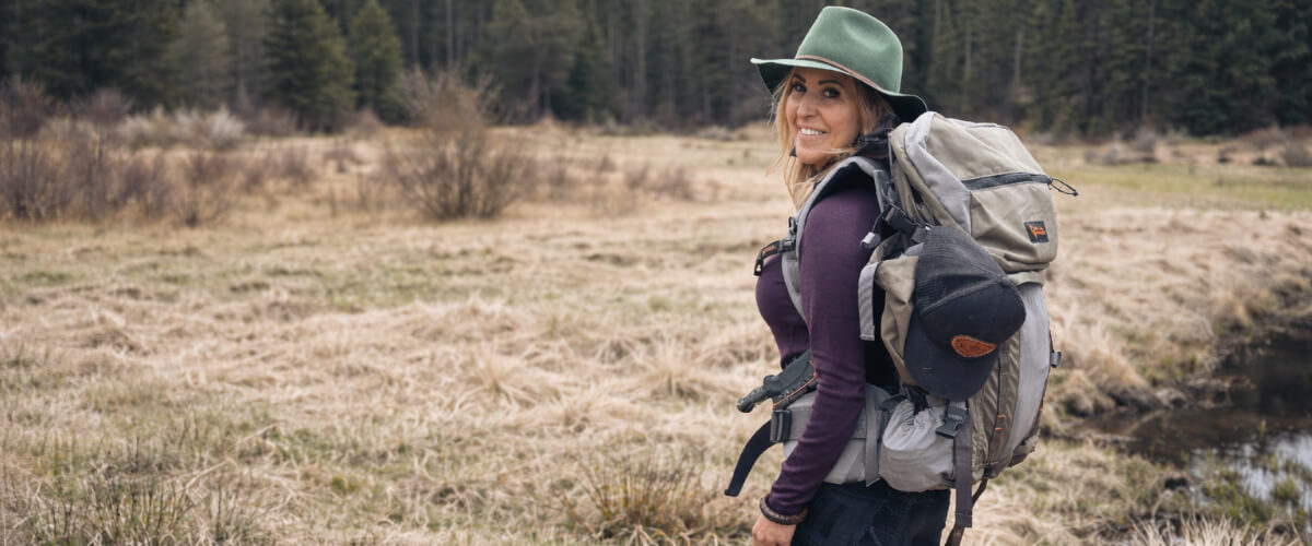 Jess Caldwell hiking solo through open meadow terrain with a backpack, demonstrating calm awareness and deliberate travel in a forested landscape.
